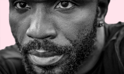 Black and White photo of a black man with a beard and mustache wearing a black t-shirt on a pink background.